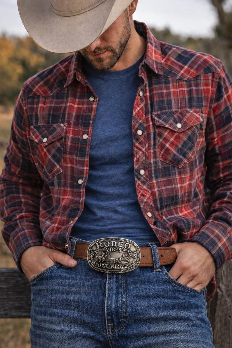 May include: A person wearing a red and navy plaid button-down shirt over a blue t-shirt, blue jeans, and a cowboy hat. A brown leather belt with a silver rodeo buckle that reads "Rodeo Vibes Love Thrives" is visible.
