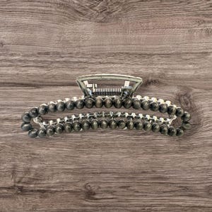 May include: A silver-toned hair claw clip with a beaded design. The clip features a rectangular top and a series of small, dark gray beads that form the main body of the clip. The clip is designed to hold hair in place.