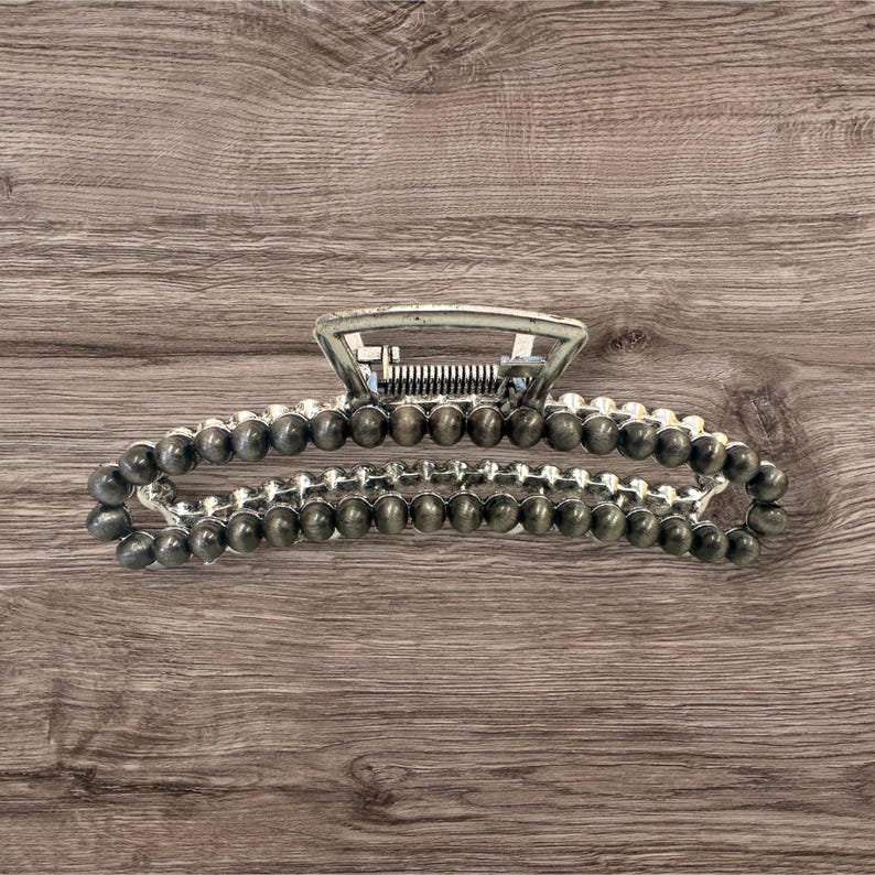 May include: A silver-toned hair claw clip with a beaded design. The clip features a rectangular top and a series of small, dark gray beads that form the main body of the clip. The clip is designed to hold hair in place.
