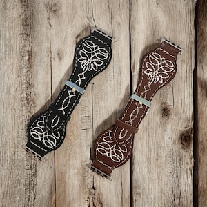 May include: Two watch bands with a western design. One band is black with white stitching, and the other is brown with white stitching. Both bands have a decorative floral pattern and a silver-colored buckle.