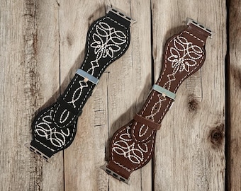 May include: Two watch bands with a western design. One band is black with white stitching, and the other is brown with white stitching. Both bands have a decorative floral pattern and a silver-colored buckle.