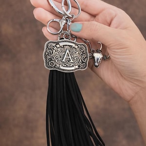 May include: Silver-tone keychain with a decorative buckle featuring the letter "A" and a black tassel. The keychain also includes a small bull head charm, perfect for adding a touch of Western flair to your keys or bag.