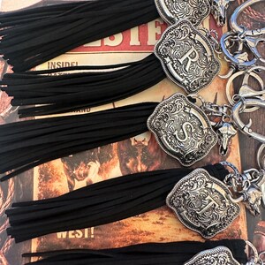 May include: Keychains with black tassels and silver-colored, engraved metal accents. Each keychain features a different letter: P, R, S, T, and W. The keychains have a Western theme, with a decorative bull skull charm.