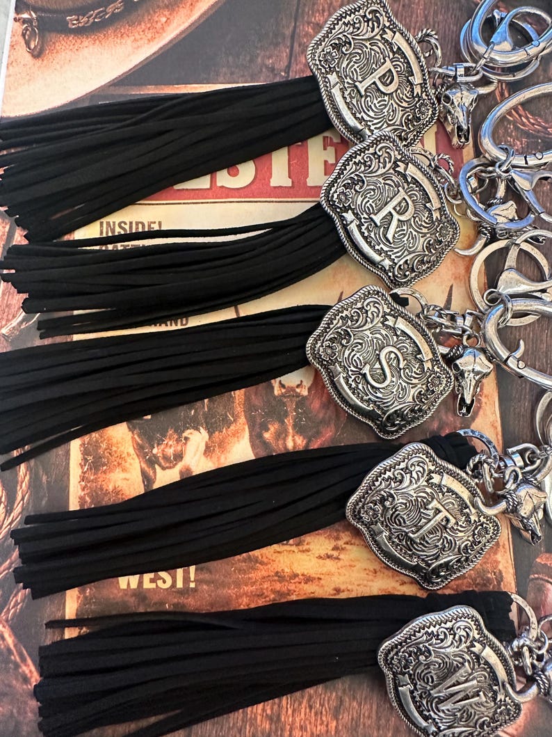 May include: Keychains with black tassels and silver-colored, engraved metal accents. Each keychain features a different letter: P, R, S, T, and W. The keychains have a Western theme, with a decorative bull skull charm.