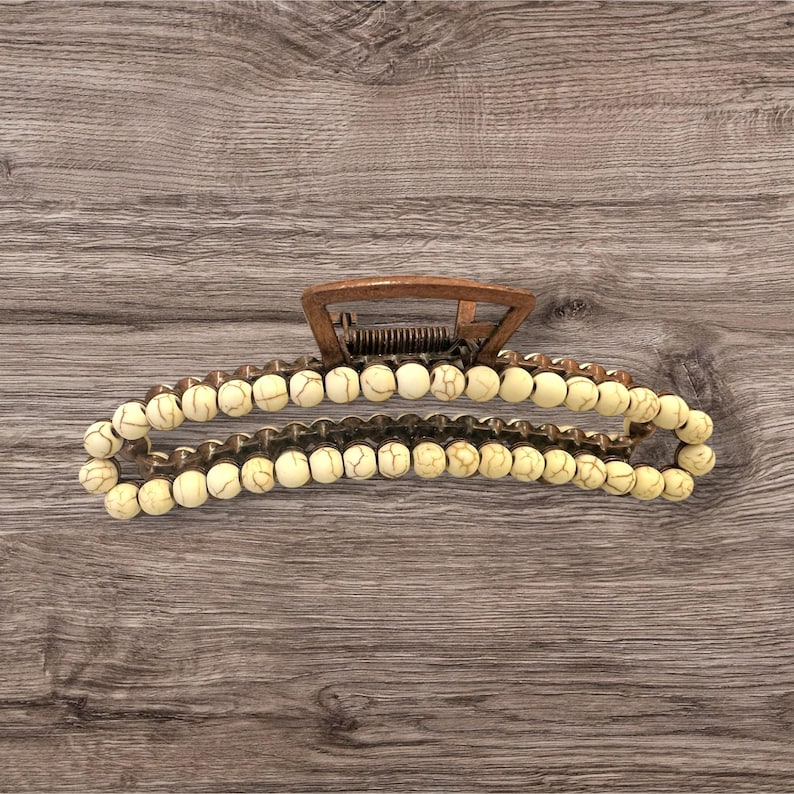 May include: A copper-colored metal hair claw clip with a row of off-white, round beads. The beads are strung along the clip's curved arms. The clip's top section has a spring mechanism for opening and closing.