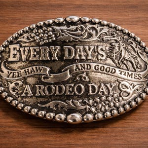 May include: Oval silver-colored belt buckle with the words "EVERY DAY'S A RODEO DAY" and "YEE HAW AND GOOD TIMES" in raised lettering. The buckle has a decorative border and is set against a wood grain background.
