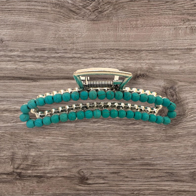 May include: A silver-toned hair claw clip with turquoise-colored beads. The clip has a hinged design with a spring mechanism and a decorative top. The beads are evenly spaced along the clip's curved arms, creating a stylish accessory.