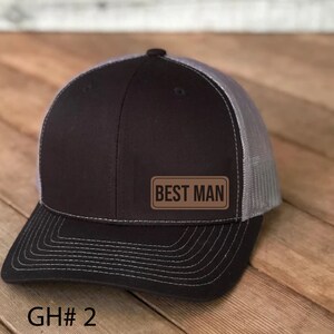 May include: A black and gray trucker hat with a brown leather patch that says "BEST MAN".