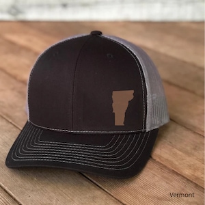 May include: A black and gray trucker hat with a brown outline of the state of Vermont on the front. The hat has a mesh back.
