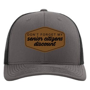 May include: Gray trucker hat with a black mesh back and a brown patch. The patch features the text "DON'T FORGET MY senior citizens discount" in black lettering. The hat has a curved brim and a snap closure.