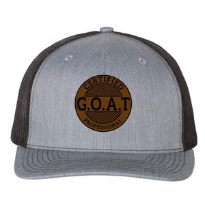 May include: A grey and black trucker hat featuring a brown circular patch on the front. The patch contains the black text "CERTIFIED G.O.A.T PROFESSIONAL" arranged in a circular pattern with diagonal line details inside the circle.