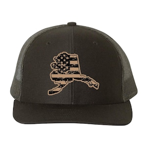 Peut inclure: Casquette de camionneur marron foncé avec un contour vieilli de l'Alaska, rempli d'un motif de drapeau américain beige et noir. La casquette a un dos en filet et une visière incurvée, idéale pour l'extérieur.