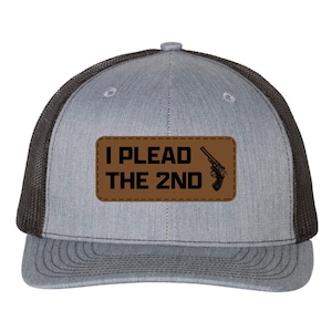 Peut inclure: Une casquette trucker grise et noire avec un écusson rectangulaire marron sur le devant. L'écusson affiche le texte noir I PLEAD THE 2ND accompagné d'un graphique de revolver. La casquette possède un panneau avant structuré et un dos en filet.