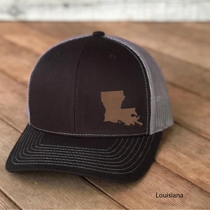 May include: A black and gray trucker hat with a brown leather patch of the state of Louisiana on the front. The text "Louisiana" is printed below the hat.