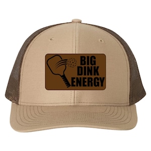 May include: A tan and brown trucker hat with a mesh back. The front features a brown rectangle with a pickleball paddle graphic and the words "BIG DINK ENERGY" in black.