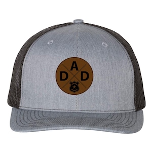 May include: A gray trucker hat with a brown leather patch featuring the word "DAD" in a cross design. The patch also includes a police badge. The hat has a black mesh back.