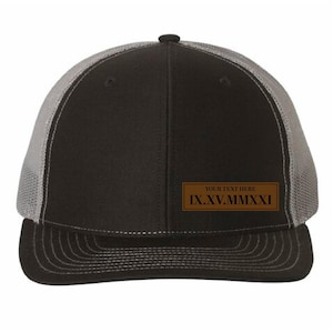 May include: Black and gray trucker hat with a curved brim. The front panel is black, and the mesh back is gray. A brown rectangular patch displays the text "YOUR TEXT HERE" and the date IX.XV.MMXXI.