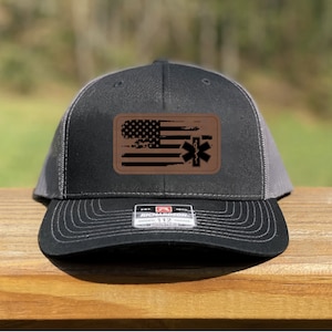 May include: A black and gray trucker hat with a brown leather patch featuring a distressed American flag with a medical symbol.