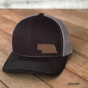 May include: A black and grey trucker hat featuring a brown leather patch of the Nebraska state outline. The hat has a curved brim and a mesh back. The word "Nebraska" is printed on the bottom right corner.