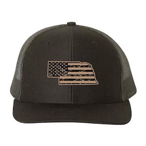 May include: A dark green trucker hat with a tan outline of the state of Nebraska and a distressed American flag design. The hat has a mesh back and a curved peak.