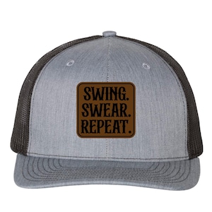 May include: A grey trucker cap with a black mesh back and a curved peak. The cap has a brown square patch with the words "SWING. SWEAR. REPEAT." in bold, dark brown letters.