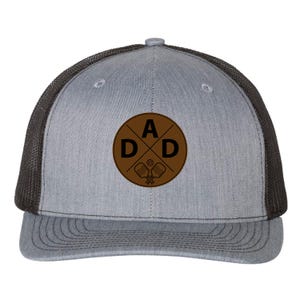 May include: Grey and black trucker hat with a brown circular patch. The patch features the word "DAD" in a cross design, with pickleball paddles and a ball. The hat has a mesh back and a curved brim.
