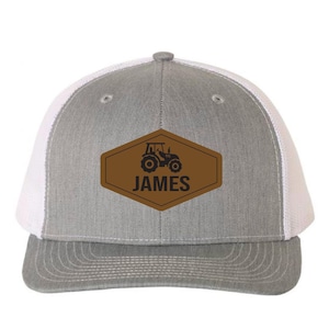 May include: Gray and white trucker hat with a brown leather patch. The patch features a black tractor graphic and the name "JAMES" in black lettering. The hat has a curved brim and a mesh back.