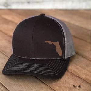 May include: A black and gray trucker hat with a brown outline of the state of Florida on the front. The hat has a mesh back and a curved brim.