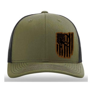 May include: Olive green trucker hat with a black mesh back and a curved brim. The front features a distressed American flag design with a silhouette of a person climbing. The hat has a snapback closure.