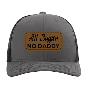 May include: A charcoal gray trucker hat with a black mesh back and a brown rectangular patch on the front. The patch features the black text "All Sugar" in a script font above the bold, capitalized black text "NO DADDY" in a sans-serif font.
