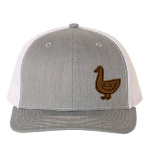 May include: A grey trucker cap with a white mesh back and a curved peak. The cap features a brown leather patch with a cartoon duck and the word "SILLY". The cap has a snap closure.