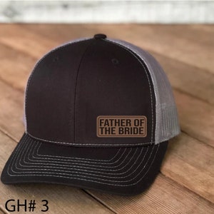 Peut inclure: Un chapeau de camionneur noir et gris avec un patch marron qui dit "Father of the Bride".