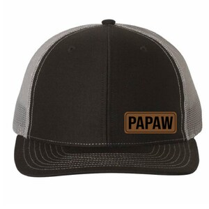May include: Black and gray trucker hat with a curved brim. The front panel is black, and the back is gray mesh. A brown rectangular patch with the word "PAPAW" in white letters is on the front.