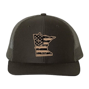 May include: A dark gray trucker hat featuring a tan outline of the state of Minnesota with a distressed American flag design. The hat has a curved brim and a mesh back.
