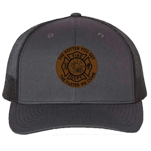 May include: A charcoal gray trucker hat with a black mesh back. The front of the hat features a brown circular patch with the words "THE HOTTER YOU GET FIRE DEPT. THE FASTER WE COME" surrounding a fire department emblem.