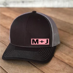 Peut inclure: Un chapeau de camionneur noir et gris avec un patch rose qui dit "M+J" dessus.