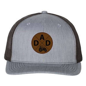 May include: Grey trucker cap with a brown circular patch displaying the word "DAD" and a tractor graphic. The cap has a grey front panel, a black mesh back, and a curved peak.