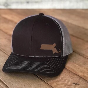 May include: A black and gray trucker hat with a brown outline of the state of Massachusetts on the front. The text "Mass." is printed below the hat.