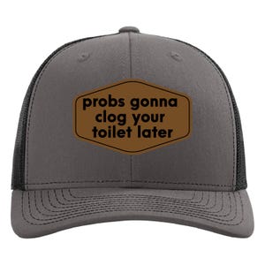 May include: A gray trucker hat with a black mesh back and a brown patch. The patch features the text "probs gonna clog your toilet later" in a playful font. The hat has a curved brim and a snapback closure.