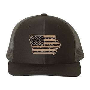 May include: A black trucker hat featuring a tan-colored patch in the shape of the state of Iowa. The patch displays a distressed American flag design with black stars and stripes against a tan background, centered on the front of the cap.