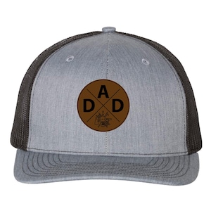 May include: A gray trucker hat with a black mesh back and a curved brim. The front features a brown circular patch with the word "DAD" and a crossed tool design.