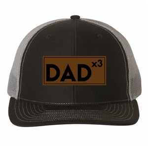 May include: A black and grey trucker hat featuring a rectangular brown patch on the front panel. The patch displays the black text DAD x3 in a bold, sans-serif font. The hat has a solid black front, a curved brim, and grey mesh side panels.