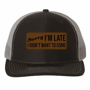 May include: Black and gray trucker hat with a brown rectangular patch. The patch features the text "Sorry I'm Late I Didn't Want to Come" in black and white. The hat has a curved brim and a mesh back.