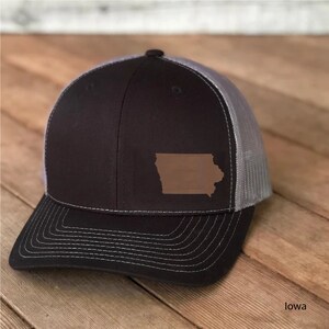 May include: A black and gray trucker hat with a brown leather patch shaped like the state of Iowa. The hat has a mesh back and a curved brim.