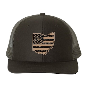 May include: A dark green trucker hat featuring a distressed design of the Ohio state outline with an American flag pattern. The hat has a mesh back and a curved brim.