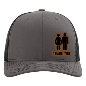 May include: A gray trucker hat with a black mesh back. The hat features a brown patch with a stick figure couple and the text "TRADE YOU" in brown letters. The hat has a curved brim.