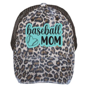 Peut inclure: Un chapeau de camionneur en tissu de type "trucker" à imprimé léopard brun et noir avec un patch bleu turquoise qui dit "baseball MOM" avec un graphique de cœur de baseball.