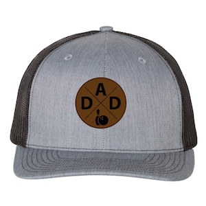 May include: Gray trucker hat with a black mesh back and a curved brim. The front features a brown circular patch with the word "DAD" in black letters and a bowling pin and ball graphic.