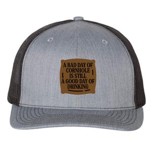 May include: Gray trucker hat with a black mesh back and a curved brim. The front features a brown patch with the text "A BAD DAY OF CORNHOLE IS STILL A GOOD DAY OF DRINKING" in black.