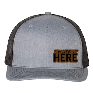 May include: A gray trucker hat with a black mesh back and a curved brim. The hat features the text "I HATE IT HERE" in a brown rectangular patch on the front.